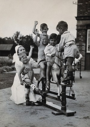 view St Nicholas' and St Martin's Orthopaedic Hospital, Pyrford, Surrey: children playing on a rocking horse with a nurse attending. Photograph, c. 1935.