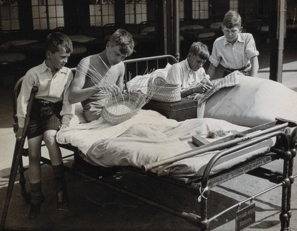 St Nicholas' and St Martin's Orthopaedic Hospital, Pyrford, Surrey: boy patients making baskets from hospital beds outdoors. Photograph, c. 1935.