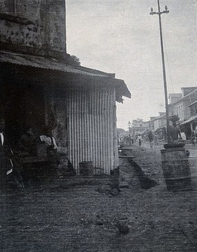 A street in Freetown, Sierra Leone: the buildings separated from the street by a ditch, with a wooden plank to connect them. Photograph, c. 1911.