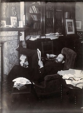 Arthur Schuster, lying on an armchair by a fireplace, with a photograph of Lord Kelvin behind him. Photograph, c. 1901.