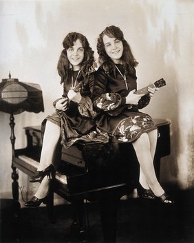 Daisy and Violet Hilton, conjoined twins, sitting on a small grand piano, holding ukeleles. Photograph, c. 1927.