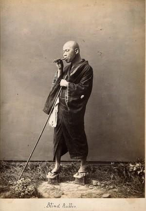 view A blind Japanese masseur with a pipe in his mouth and a long pole in his hand. Photograph.