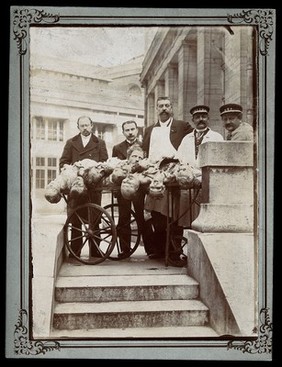 Four men wearing aprons and uniforms standing by a trolley with half-dissected cadavers on it. Photograph.