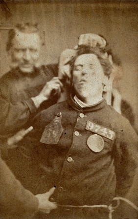 Wakefield Prison in Yorkshire: a young man, prisoner number 9743, struggling against two warders, one in front of and one behind him; head and shoulders. Photograph, c. 1869.