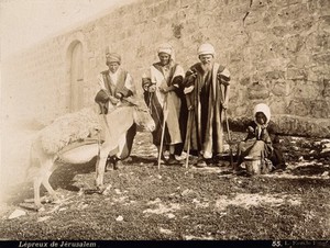 view A group of people with leprosy in Jerusalem: three standing men with sticks, a woman crouching next to a small bucket, and a mule. Photograph.