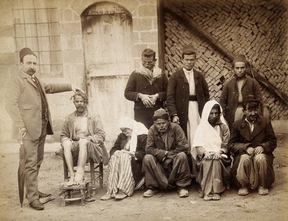 A group of people with leprosy in Jerusalem: a man in a striped suit leaning on an umbrella has his hand on the head of one of them. Photograph.