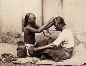 view Two Indian men sitting cross-legged; one appears to be a barber and is shaving the front of the other's head, presumably to match his own hairstyle. Photograph.