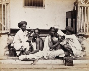 view Two Indian barbers wearing turbans shaving the heads of two Indian men who are sitting back to back on some stone steps. Photograph.