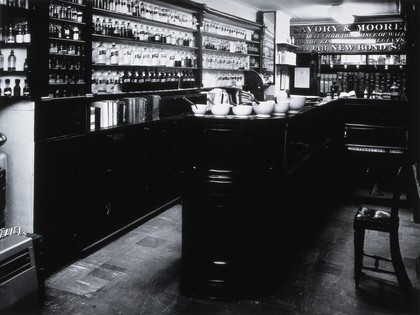 Savory & Moore Ltd, London: interior of the pharmacy with a long wooden bench and shelves of labelled bottles and jars holding drugs. Photograph.