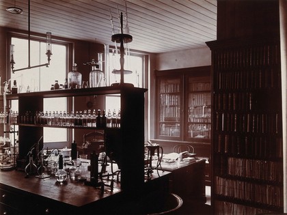 The interior of a pharmaceutical or chemical laboratory with a bench in the centre and books and papers on shelves around the walls. Photograph.