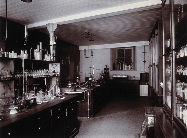 The interior of a pharmaceutical or chemical laboratory, with two long benches in the centre and glass-fronted cabinets along the right wall. Photograph.