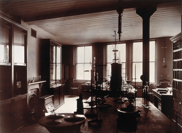 The interior of a pharmaceutical or chemical laboratory, with two long benches in the centre, glass-fronted cabinets and a coal-burner along the left wall. Photograph.