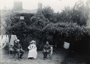 view Holmleigh Auxiliary Military Hospital, Harrow: a nurse and two soldiers, one of whom has a bandage on his head, sitting on deck chairs in a garden with blossom trees in the background. Photograph, c. 1922.