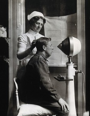 view A young uniformed man sitting with a pillow on his chair, facing a machine giving electro-magnetic treatment; a nurse holds his head in position. Photograph.