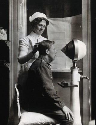 A young uniformed man sitting with a pillow on his chair, facing a machine giving electro-magnetic treatment; a nurse holds his head in position. Photograph.
