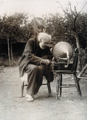 Isaac Lamb, a centenarian, sitting on a chair outdoors, winding the handle of a gramophone. Photograph, ca 1921.