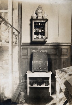 view Some objects from the Musée Pharmacie: a small cabinet with pharmacy bottles and jars and a wooden box decorated with metal. Photograph.