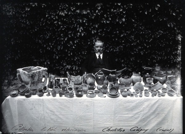 A man with long moustaches (E. Viol?) seated behind a white clothed table which is filled with pharmacy jars and other objects from the collection of E. Viol. Photograph.