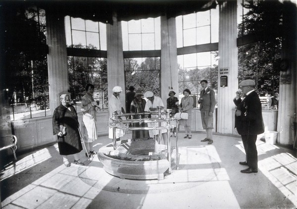 Franzensbad (Františkovy Lázně), Czechoslovakia: the interior of the Francis Spring where people stand to drink water drawn from the central circular point by female attendants in white uniforms and caps. Photograph.