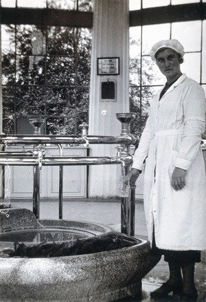 view Franzensbad (Františkovy Lázně), Czechoslovakia: a female attendant in the building which houses the Francis Spring, filling a glass with water from a tap attached to circular pipes leading to the central source. Photograph.