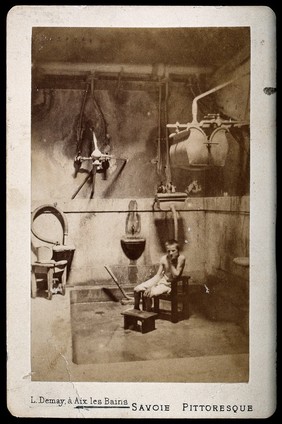 A boy sitting on a wooden chair, his chin resting on his hand, in a room used as a "shower bath" at Aix les Bains. Photograph by L. Demay.