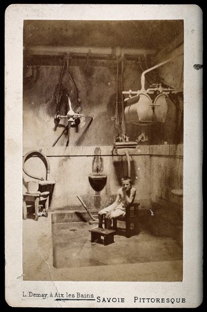 view A boy sitting on a wooden chair, his chin resting on his hand, in a room used as a "shower bath" at Aix les Bains. Photograph by L. Demay.