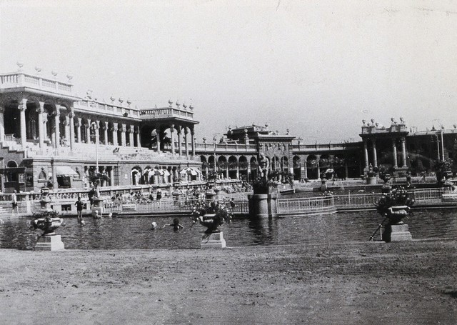 Open air baths in Budapest; two large pools separated by railings in which people are swimming. Photograph.