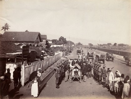 A group of people in Bombay, mostly uniformed, standing around a wooden bed marked 17 on which a man is lying, during an outbreak of plague. Photograph, 1896/1897.
