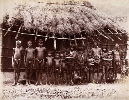 A group of adults and children standing in a line outside a bamboo hut with a thatched roof: Bombay at the time of the plague. Photograph, 1896/1897.