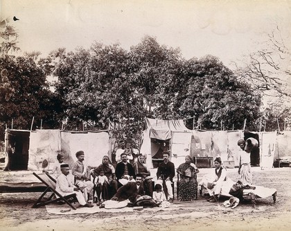 An affluent family forced to leave their home due to plague in their neighbourhood sitting outside temporary huts in a camp: Bombay at the time of the plague. Photograph, 1896/1897.