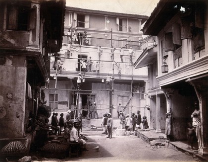A plague house being whitewashed by men standing on scaffolding in Bombay. Photograph, 1896.