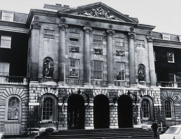 The front entrance of Guy's Hospital, with statues of Aesculapius and Hygieia above the arches. Photograph.