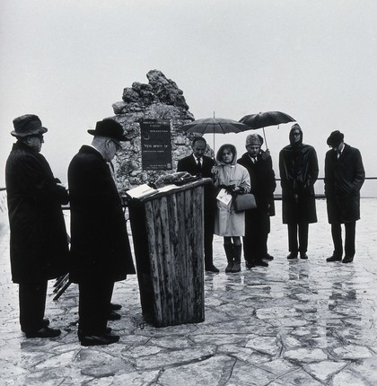 Rabbi Dr. Philipp and Reverend Lipschitz addressing a group of spectators, in the rain, adjacent to a memorial in Israel to Thomas Guy, founder of Guy's Hospital. Photograph.
