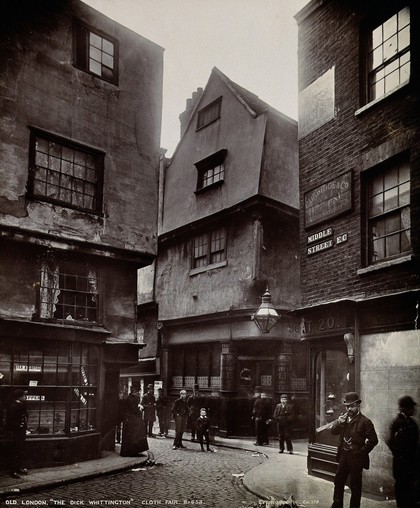 The church of St. Bartholomew the Great and surrounding area; the Dick Whittington pub on the corner of a street. Photograph.