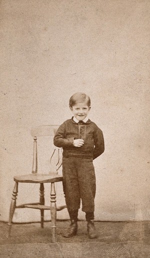 view A boy smiling and standing next to a wooden chair. Photograph.