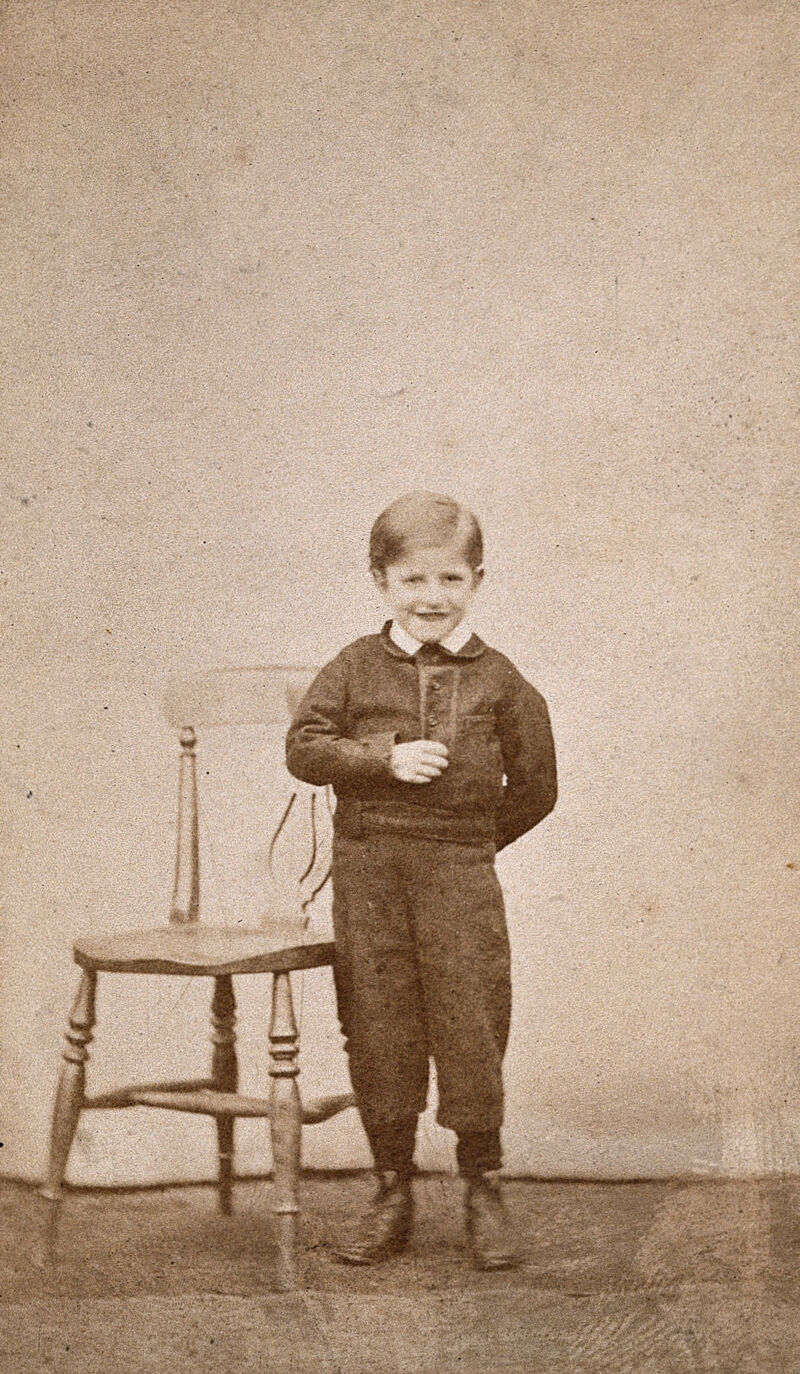 A boy smiling and standing next to a wooden chair. Photograph., by John Davis