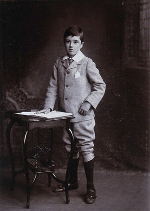 view A young man, formally dressed, resting his hand upon an open book upon a table. Photograph by Lock & Whitfield.