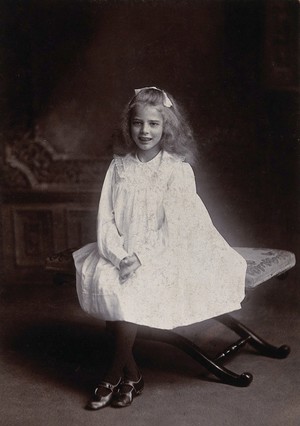 view A girl wearing a white dress, sitting on an embroidered stool. Photograph by Lock & Whitfield.