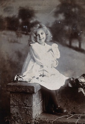 A girl, holding a doll, sitting on a wall. Photograph by Lock & Whitfield.