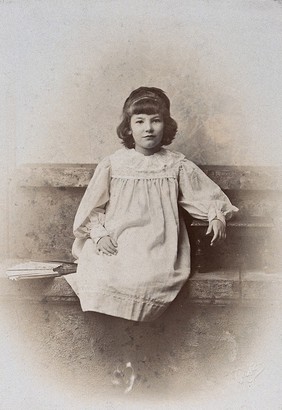 A girl, sitting on a stone bench with a fan folded at her side. Photograph by A. Reston.