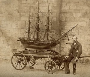 view James Pullen, sitting on the end of a wooden trolley upon which rests a huge model ship made by him. Process print.