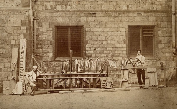 Two men standing next to a complex wooden apparatus. Process print.