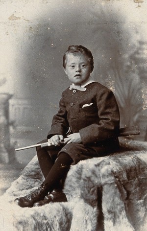 view A boy with Down's syndrome, holding a rifle and sitting on a fur carpet. Photograph by R.W. Morris.