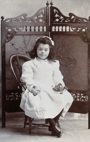 view A girl with Down's syndrome, sitting in front of an ornate wooden screen, holding a small posy of flowers. Photograph by Chas Marshall.