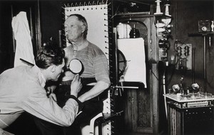 view Radiography: a seated man is attached to equipment by a technician, to have an x-ray taken. Photograph, ca. 1925.