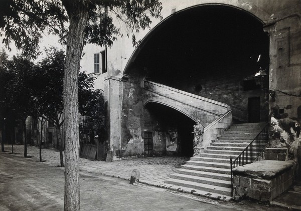 Hospital de la Santa Cruz, Barcelona: a staircase leading up to the men's department; at the foot there is a statue of Charity. Photograph.