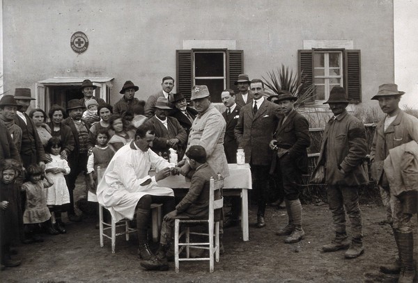 Nettuno (?), Italy: a medical man taking a blood sample from a boy seated at a small table, surrounded by a crowd of onlookers, outside the local ambulance service building (?). Photograph, 1918/1937 (?).