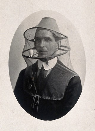 Mosquito headnet with cap, modelled by a man smoking a pipe. Photograph, 1902/1918 (?).
