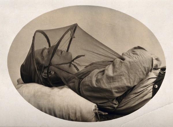 Mosquito headnet with armholes, covering upper body, modelled by a sleeping man. Photograph, 1902/1918 (?).