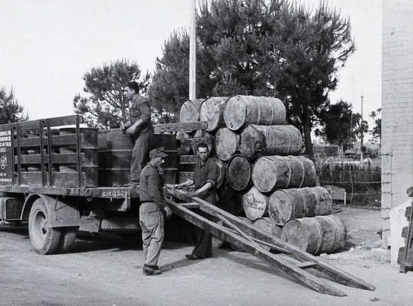 Malaria control supplies, Italy: large metal barrels being unloaded from a truck by three men. Photograph, 1940/1950 (?).